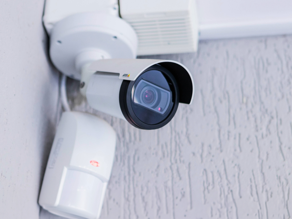 Câmera de segurança Bullet Axis instalada em parede com sensor de alarme integrado. O modelo possui lente de alto desempenho, protegida por uma carenagem para uso externo. O sensor de alarme PIR instalado abaixo da câmera detecta movimento e pode acionar gravação ou alertas de segurança em tempo real. Essa configuração é ideal para sistemas de CFTV inteligentes, combinando monitoramento por vídeo e detecção de presença para maior proteção.