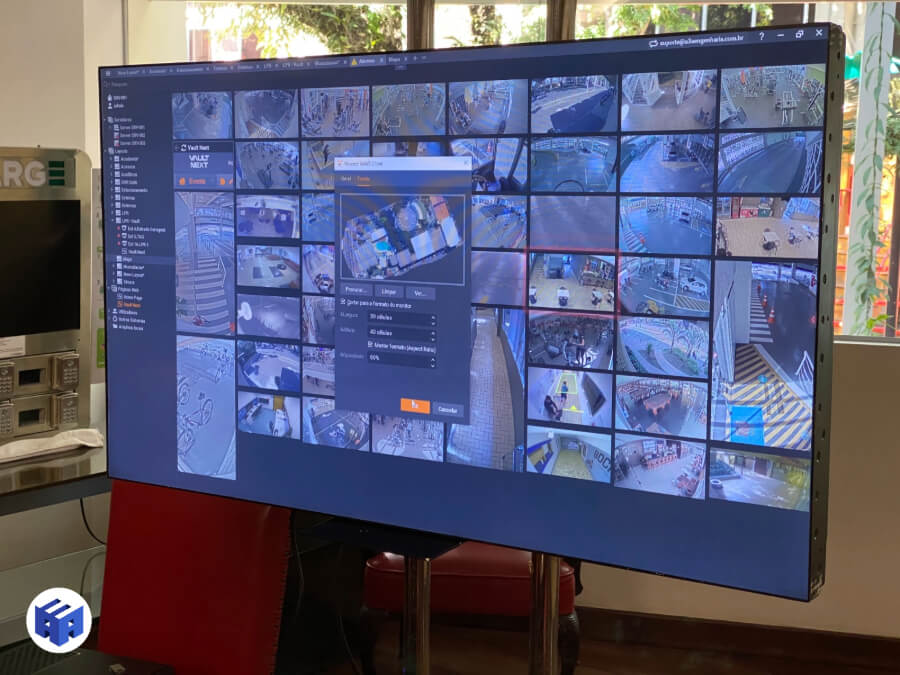 Interface gráfica de um software VMS (Video Management System) exibida em um monitor grande, apresentando múltiplos feeds de câmeras de segurança em um ambiente corporativo. A tela mostra uma grade de visualização com diversas câmeras monitorando diferentes áreas, incluindo corredores, entradas e espaços internos. No centro, uma janela de configuração está aberta, permitindo ajustes no armazenamento e qualidade de gravação. Ideal para controle centralizado de CFTV, monitoramento remoto e gestão de segurança.